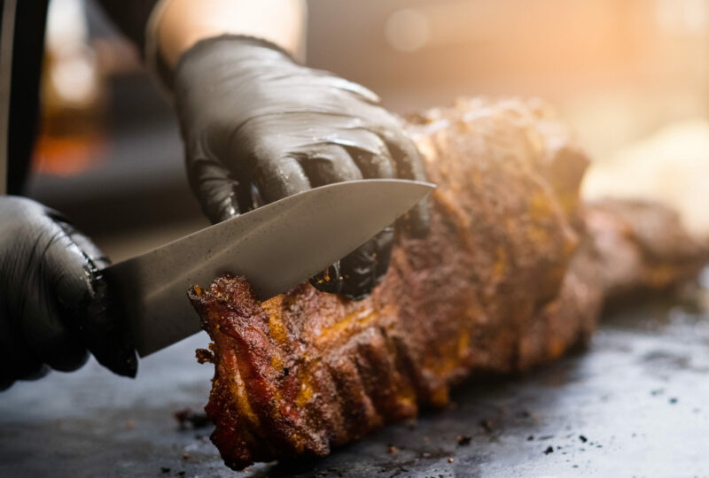 Grill restaurant kitchen. Chef in black cooking gloves using knife to cut smoked pork ribs.
